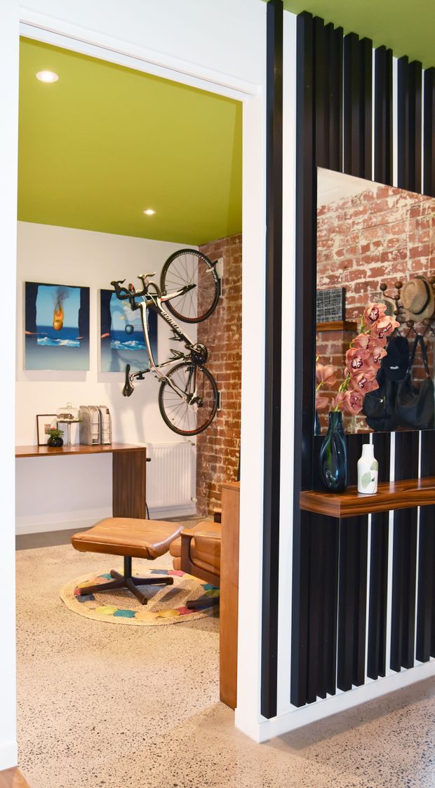 A colorful eclectic interior view looking into a compact living or lounge area. The room features a vivid lime-green ceiling, exposed red brick walls, polished speckled concrete or terrazzo-like flooring, a wall-mounted bicycle, framed artwork, a mid-century-style leather ottoman and chair, and a dark vertical slat feature wall with a timber display shelf and decorative objects.
