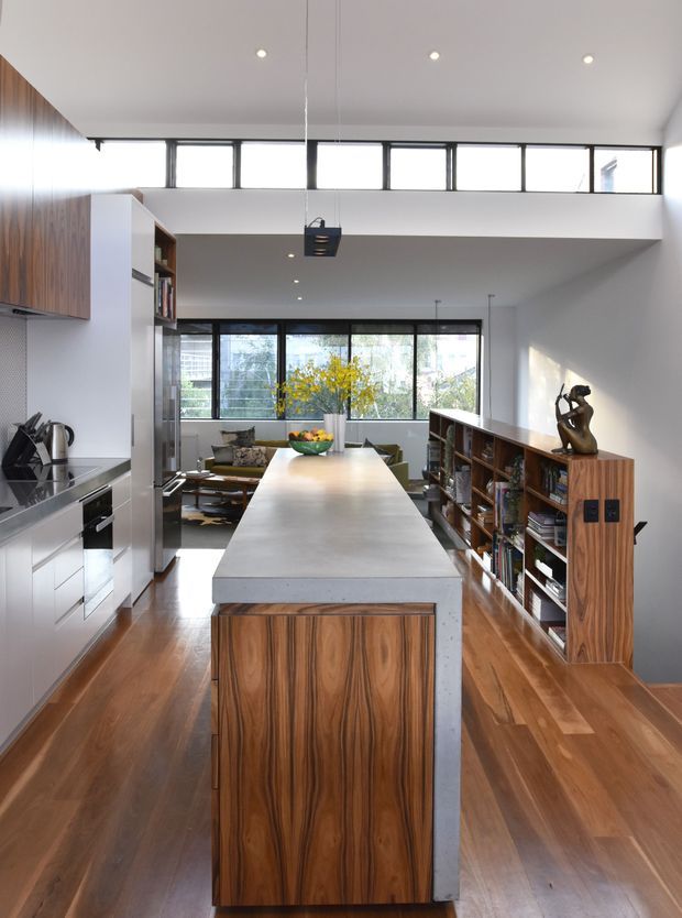 A bright, open-plan contemporary kitchen viewed from the kitchen island, with a long concrete island benchtop and timber-veneered ends, white cabinetry, stainless-steel appliances, and warm timber flooring. A large built-in timber bookshelf divides the kitchen from the living area beyond. High clerestory windows, extensive glazing, recessed ceiling lights, and a suspended pendant create an airy, high-volume interior.