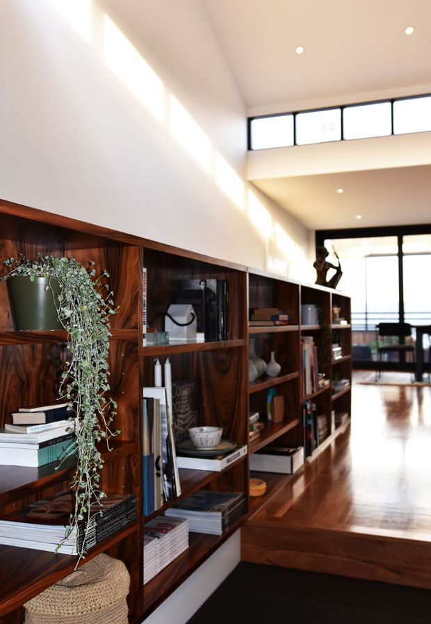 A bright, double-height residential interior featuring a long wall of dark timber built-in shelving and cabinetry filled with books and decorative objects. A trailing plant adds greenery in the foreground. Polished timber flooring leads toward a glazed living or dining area, with clerestory windows, recessed ceiling lights, and a sculptural figure visible near the far end.