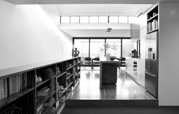 Monochromatic photograph of a bright, open-plan contemporary kitchen viewed from a lower living or circulation area. The kitchen features a central island, extensive integrated cabinetry and shelving, stainless-steel appliances, polished timber flooring, high clerestory windows, recessed lighting, and large glazed doors opening to an exterior terrace or balcony. A step-up transition separates the foreground from the kitchen.