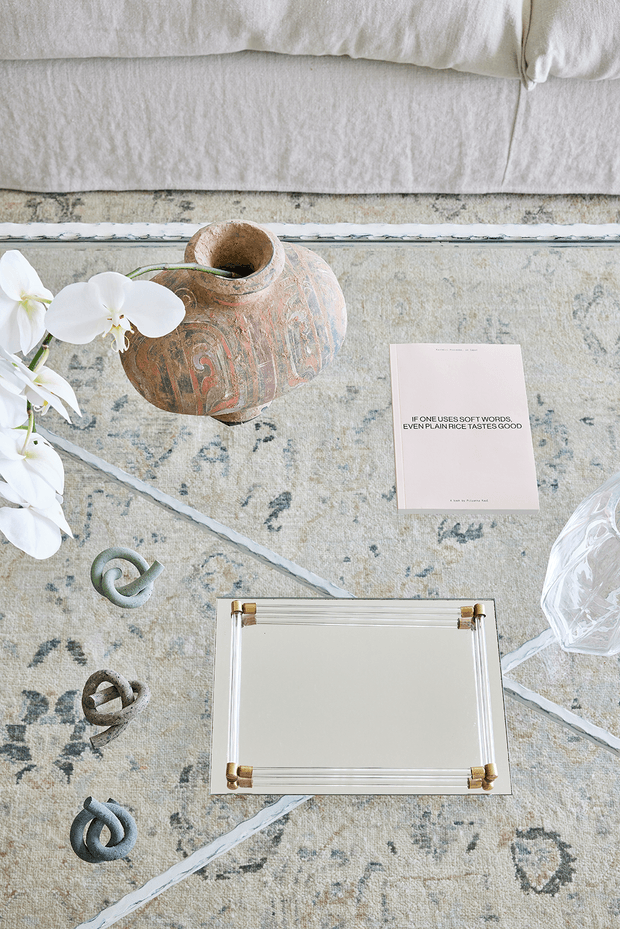 Overhead still-life composition on a pale patterned rug beside a light upholstered sofa. The arrangement includes a weathered terracotta vessel with white flowers, a small printed card, sculptural looped objects, a clear faceted vessel, and a rectangular mirrored tray with brass corner details. The image has a soft, refined, neutral palette with decorative styling rather than a clearly identifiable room.