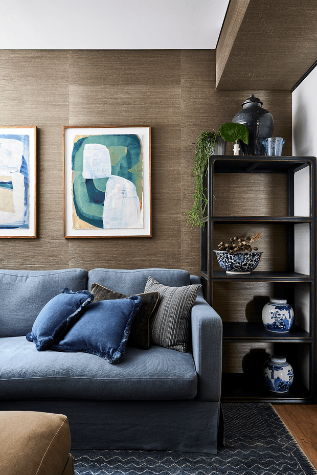 A refined living room vignette featuring a deep blue upholstered sofa with layered blue and striped cushions, set against textured taupe-brown wall panelling. A black open display shelving unit holds blue-and-white ceramics, glassware, a large dark vessel, and trailing greenery. Abstract framed artwork, a dark patterned rug, timber flooring, and a partial tan ottoman complete the layered, sophisticated interior.