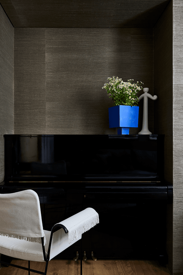 A moody, refined music nook featuring a glossy black upright piano set into a dark textured wall alcove. A white fringed lounge chair sits in front of the piano, while a vivid blue planter with delicate flowers and a sculptural white decorative object provide accents. Warm timber flooring contrasts with the dark walls and piano.
