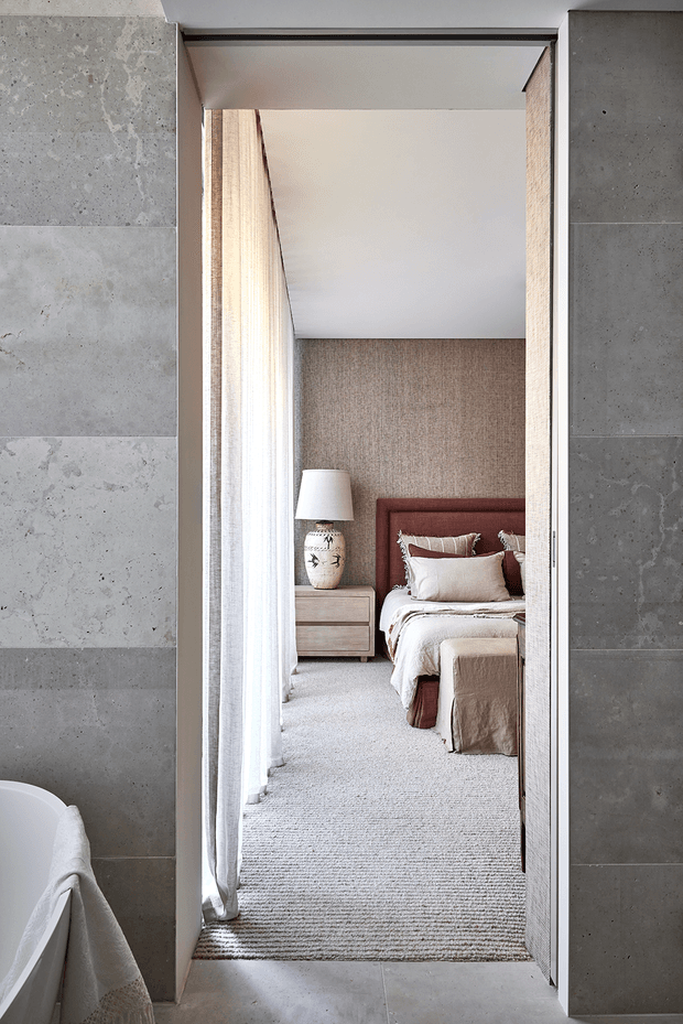 A refined view through a wide doorway from a tiled bathroom into a softly styled bedroom. The bathroom features large grey stone-look wall and floor tiles with the edge of a freestanding white bathtub visible in the foreground. Beyond the opening is a warm, neutral bedroom with textured beige wall covering, a rust-toned upholstered bedhead, layered cream and taupe bedding, bedside furniture, sheer curtains, and a pale textured carpet. The composition emphasises the connection between the bathroom and bedroom.