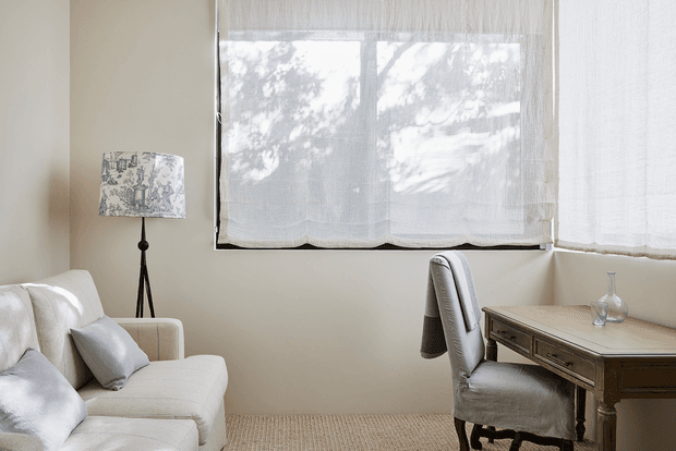 A softly lit, neutral-toned room combining a small living area with a traditional writing desk. An upholstered cream sofa sits beside a patterned floor lamp, while a cushioned desk chair and dark timber desk occupy the corner by sheer window coverings. The room has a calm, understated and refined atmosphere, with woven carpet flooring and warm natural light.