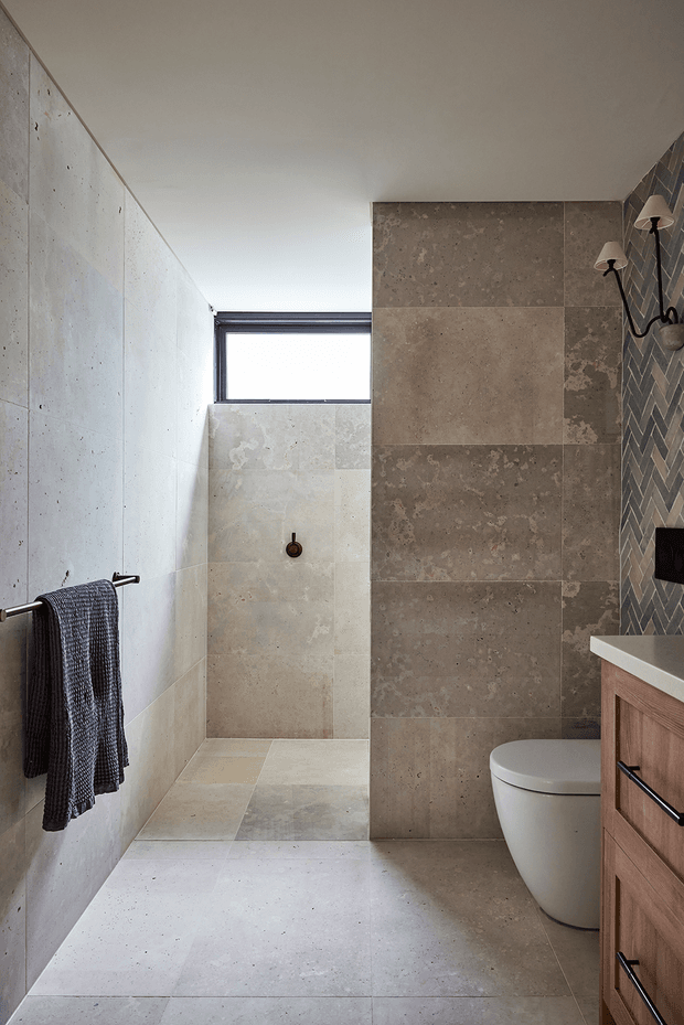 A contemporary bathroom featuring a walk-in tiled shower at the rear, a high horizontal window, wall-hung-style toilet, timber vanity with a white benchtop, and dark metal wall sconces. Large warm grey-beige stone-look tiles cover the walls and floor, with a darker patterned accent tile beside the vanity.