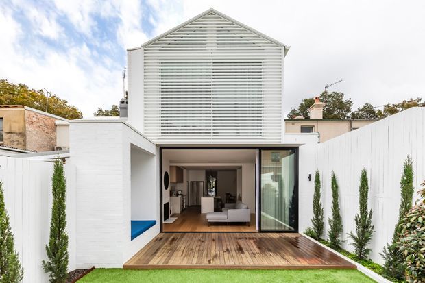 Rear exterior view of a narrow, contemporary two-storey residence with a prominent gabled upper facade screened by horizontal white louvres. The ground level opens onto a timber deck and compact lawn, with a living room visible through wide glazed sliding doors. White rendered or painted masonry walls, privacy fencing, slender planted borders, and a blue-lined recessed outdoor feature contribute to the clean courtyard-like composition.