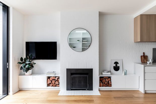 Bright open-plan living area with a white-painted brick fireplace wall, black built-in fireplace, wall-mounted television, low white media cabinetry, open firewood storage, and warm timber flooring. A compact white kitchen with wood-toned upper cabinetry is visible to the right. Floor-to-ceiling glazing at left brings in abundant natural light, while a round wall mirror and indoor plant add visual interest.