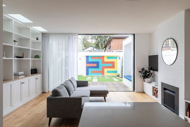 Bright contemporary living room with a grey sectional sofa, pale timber flooring, extensive white built-in shelving and cabinetry, and a white-painted brick fireplace wall. Large sliding glass doors open onto a timber deck and a colourful geometric outdoor feature wall, with a compact garden courtyard beyond.