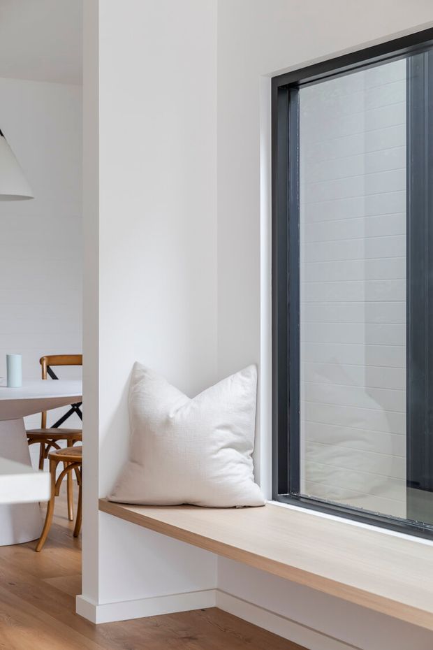 Bright minimalist interior detail showing a built-in window seat with a white cushion beneath a large dark-framed window. The adjoining dining area is visible to the left, with a light timber floor, white walls, and timber dining furniture. The space has clean lines, restrained decoration, and abundant natural light.