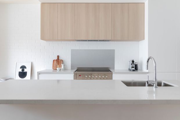 Bright, minimalist kitchen viewed across a large island. The space features pale timber overhead cabinetry, white textured brick-look wall tiles, a white island benchtop, integrated cooktop and oven, stainless-steel sink and gooseneck tap, and a compact coffee machine. The restrained palette and uncluttered composition create a clean contemporary feel.