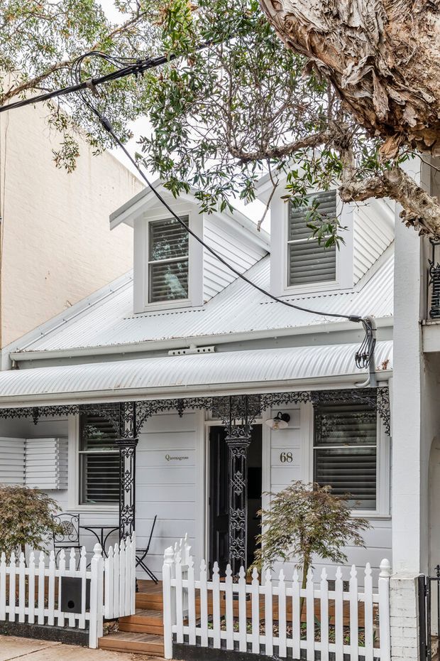 Street-facing view of a compact heritage-style cottage facade with white weatherboard cladding, a corrugated metal roof, dormer windows, decorative black wrought-iron veranda detailing, timber steps, and a white picket fence. The home is partially framed by a mature tree and overhead service wires.