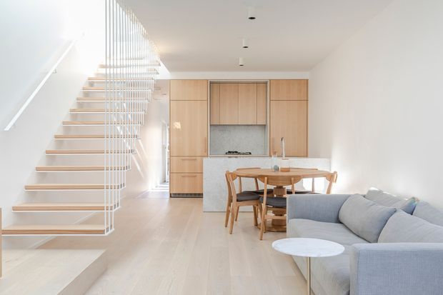 Bright, open-plan contemporary interior combining a compact kitchen, dining area and living room. Pale timber cabinetry and flooring create a warm neutral backdrop, while a white sculptural open-riser staircase with vertical balustrade occupies the left side. A light grey sectional sofa and round dining table define the living and dining zones.