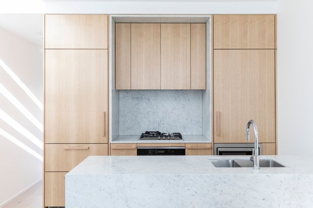 Bright contemporary kitchen with a full-height light timber cabinetry wall, integrated appliances, a marble-look stone splashback and large island bench with an undermount sink and tall chrome tap. Strong sunlight creates diagonal highlights on the pale wall.