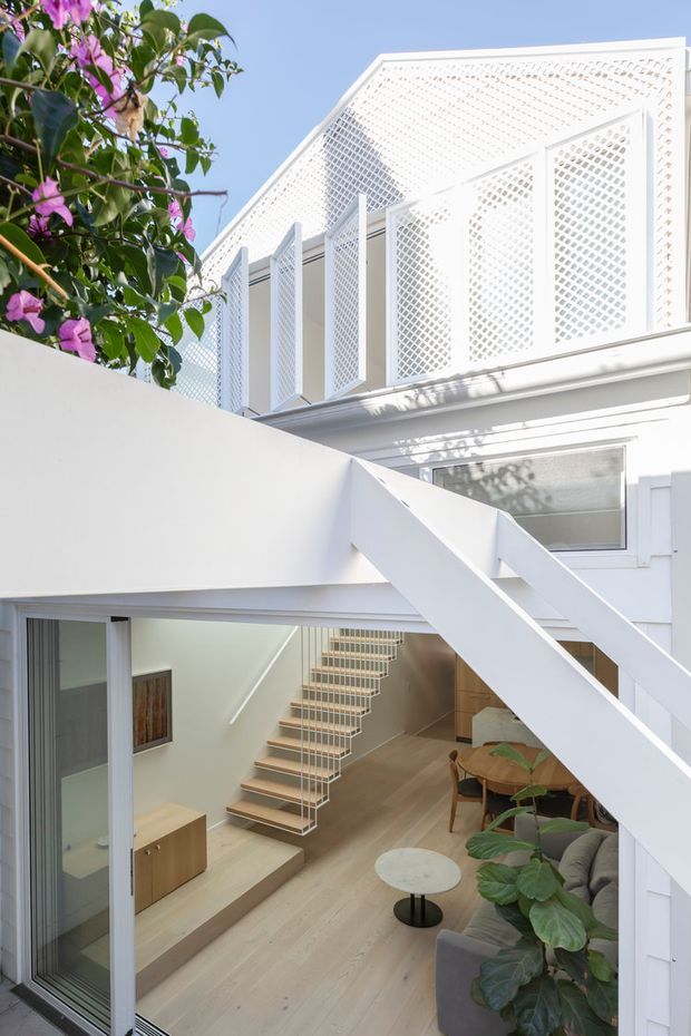 A bright, multi-level residential interior viewed from an open courtyard or stair landing. White-painted walls and structural elements frame a floating timber staircase with slim metal balustrades, leading to an upper level enclosed by decorative white lattice screening. The lower level contains a contemporary living and dining area with a grey sofa, round coffee table, timber dining table, built-in cabinetry, pale timber flooring, and large glazed doors. Tropical greenery and pink bougainvillea appear along the courtyard edge. The composition highlights indoor-outdoor connection, layered circulation, and abundant natural light.