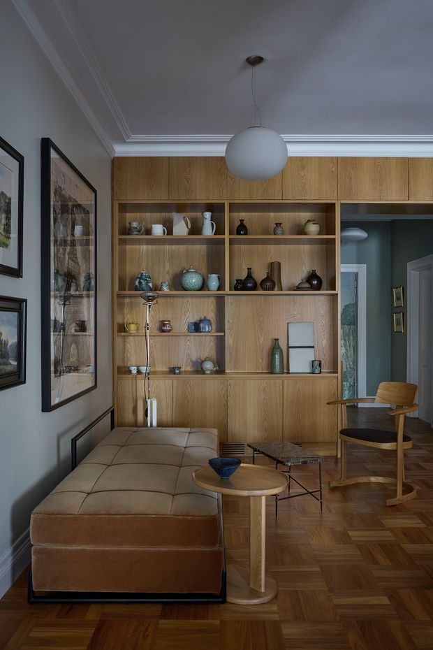 A refined living room with a warm mid-century-inspired interior. The room features extensive built-in timber shelving and cabinetry, a tan upholstered daybed or sectional, curved wood seating, nesting coffee tables, a spherical pendant light, framed artwork, and richly patterned parquet timber flooring. Decorative ceramics fill the open shelving, while pale blue-grey walls, ornate ceiling cornices, and dark trim create a sophisticated, layered atmosphere.