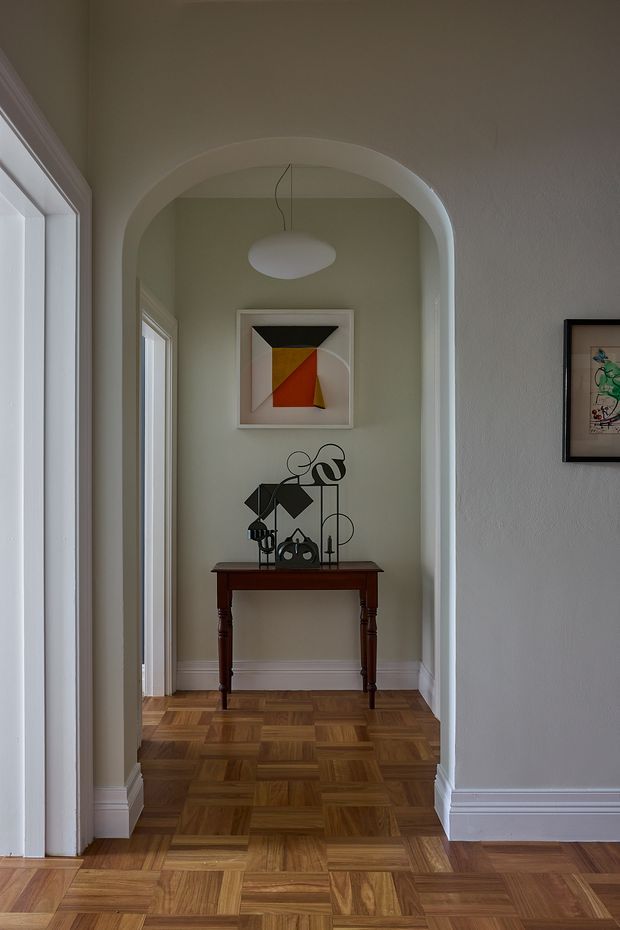 A narrow, refined hallway or entry passage framed by a large rounded archway. The space has pale grey-green textured walls, white decorative skirting and trim, warm parquet timber flooring, and tall white-framed windows or glazing on the left. A dark wood console table with turned legs sits beneath abstract artwork and a sculptural black metal object. A simple white pendant hangs within the arched recess, creating a calm, gallery-like interior.