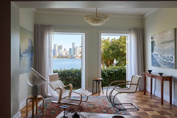 A refined living room with parquet flooring, pale walls, sheer curtains, and large windows overlooking a waterfront skyline. The room features sculptural mid-century-style lounge chairs, a patterned rug, a crystal chandelier, a timber console, and coastal artwork.
