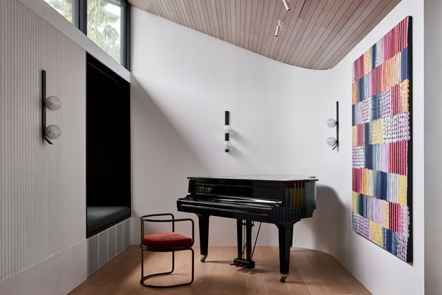 Refined contemporary music room featuring a glossy black grand piano, a sculptural black metal chair with a red upholstered seat, white curved walls, warm timber slatted ceiling, timber flooring, minimal wall sconces, and a large colourful woven wall artwork. Built-in vertical fluted wall panelling and high windows add architectural interest.