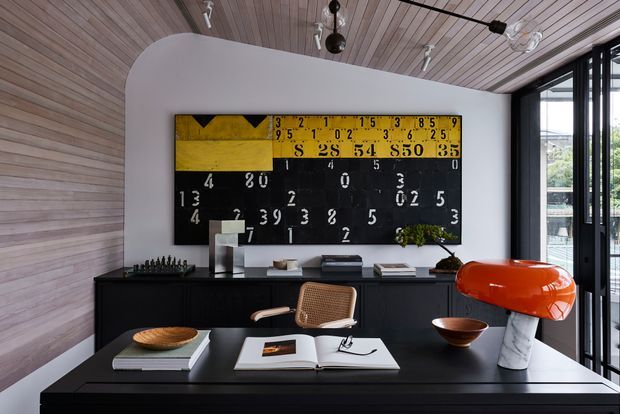 A refined contemporary living area with a dramatic black built-in credenza and large black worktable, styled with books, ceramics, a woven chair, and an orange sculptural lamp. The room features a curved white partition wall, vertical timber wall and ceiling lining, black-framed glazing, and a bold vintage-inspired black-and-yellow artwork.