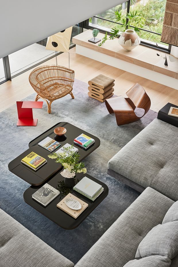 A sophisticated contemporary living room viewed from above, featuring expansive floor-to-ceiling glazing, pale timber flooring, a built-in timber window seat, a large grey rug, and a grey sectional sofa. Sculptural furniture includes a woven rattan lounge chair, a curved wooden lounge chair, stacked sculptural stools, black rounded coffee tables, and a bold red side table. Neutral earthy finishes are complemented by indoor plants, artwork, books, and decorative objects.