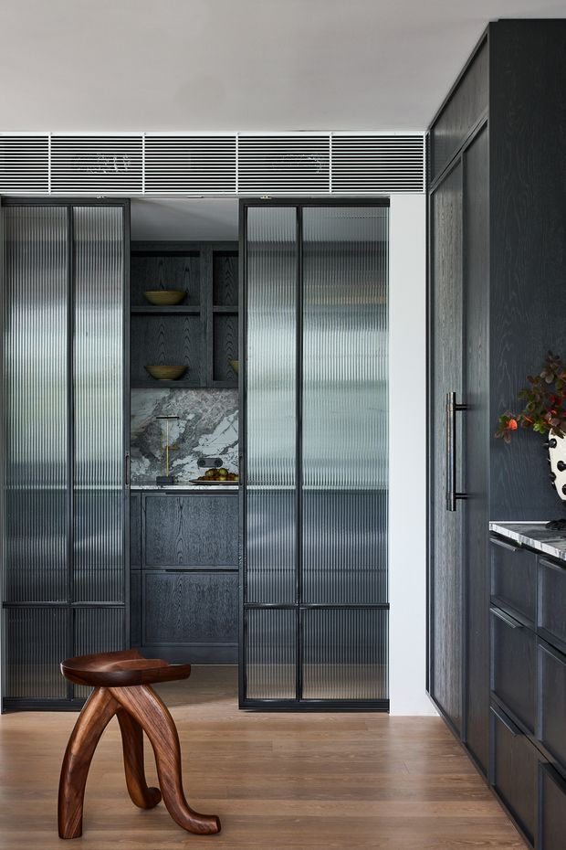 A refined dark-toned kitchen or pantry area with full-height ribbed translucent glass sliding doors in black frames, dark timber cabinetry, and a dramatic marble backsplash and benchtop. Open shelving displays bowls and produce, while warm timber flooring and a sculptural wooden stool add contrast. The space has a tailored contemporary feel with strong vertical and horizontal lines.
