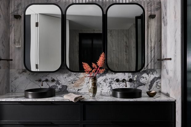 A dramatic, symmetrical bathroom vanity with two black vessel basins set on a dark built-in cabinet. The wall features richly veined marble, three rounded-rectangle black-framed mirrors, black wall-mounted tapware, and vertical grey timber-like panelling reflected in the mirrors. A small vase with orange foliage provides a warm accent.