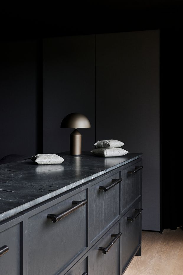 Moody close-up of a dark kitchen island or built-in cabinetry with a thick black stone benchtop, charcoal panelled drawers and doors, and dark metal bar pulls. A small sculptural table lamp and pale decorative objects sit on the benchtop. The space has dark walls and warm light timber flooring visible at the edge.