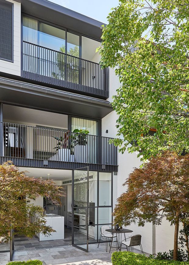 A contemporary multi-level residential facade enclosing a landscaped courtyard. The building features white rendered walls, dark metal-framed glazing, slim black balcony balustrades, and a ground-level outdoor dining area beside a glazed entry or courtyard enclosure. Layered Japanese maple-style planting softens the clean architectural lines.