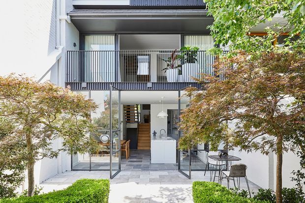 A bright contemporary multi-storey residential facade viewed from a landscaped courtyard. The lower level opens through large glazed doors to a compact kitchen and dining area, while the upper level has a balcony with dark vertical metal balustrades and extensive glazing. White rendered walls, grey stone-like paving, clipped hedges, and Japanese maple trees create a clean, refined courtyard setting.