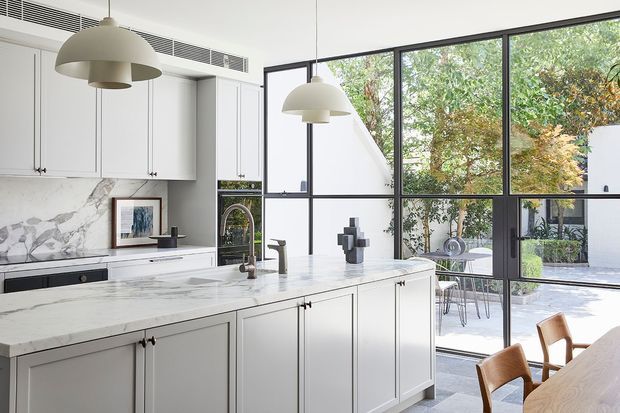 Bright contemporary kitchen with a large marble island, white shaker-style cabinetry, black-framed floor-to-ceiling glazing, and pendant lights. The glazing opens visually to a leafy courtyard or patio, while a dining table and chairs are visible in the foreground.