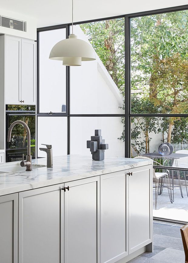 Bright contemporary kitchen with white shaker-style cabinetry, a pale veined stone island benchtop, dark-framed floor-to-ceiling glazing, and a sculptural pendant light. The kitchen opens to a leafy outdoor terrace with a small dining setting.