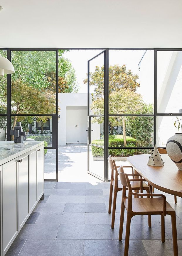 Bright contemporary open-plan kitchen and dining area opening onto a landscaped courtyard. The space features a large white island with dark hardware, pale stone or porcelain tiled flooring, black-framed floor-to-ceiling glazing and an open glass door, plus a timber dining table and matching wooden chairs. The courtyard and leafy planting are clearly visible beyond the glazing.