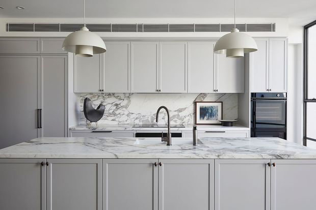 Bright, refined contemporary kitchen viewed directly across a large marble island. The room features pale grey shaker-style cabinetry, a matching marble benchtop and splashback, integrated appliances, a dark metal tap, and two oversized white pendant lights. A full-width linear air-conditioning grille and large window contribute to the clean, tailored fit-out.