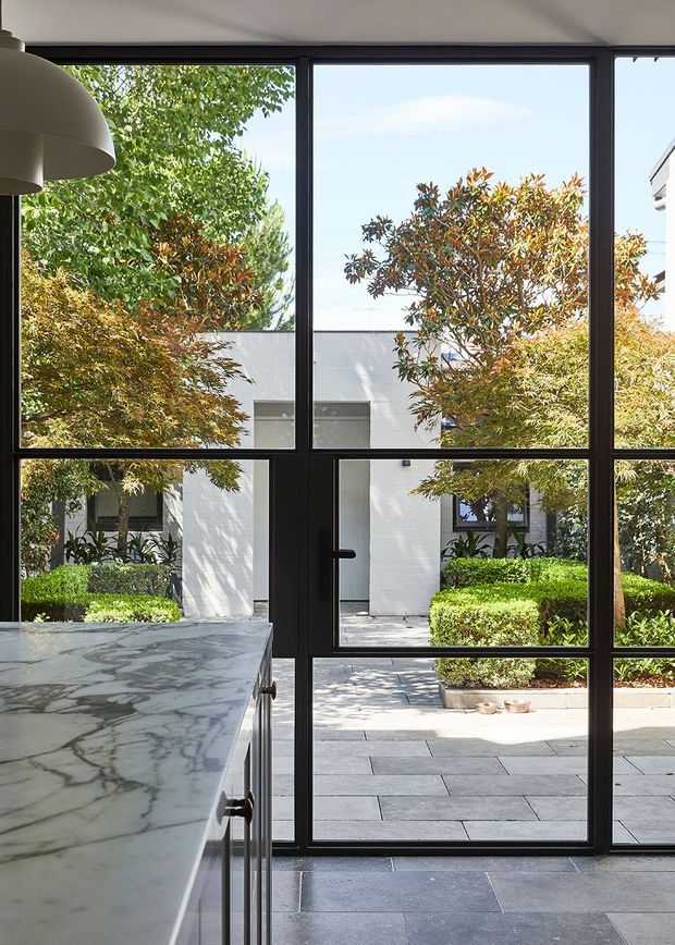 A refined interior view through expansive black-framed steel or aluminium glazing and a glazed door into a landscaped courtyard. A substantial white veined stone kitchen island occupies the foreground, with dark large-format flooring and a simple curved pendant light partially visible overhead. The courtyard features pale paving, clipped hedges, mature leafy planting, and a white rendered or masonry building beyond.