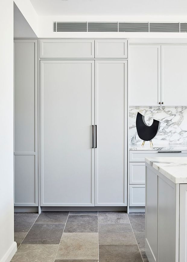A bright, refined kitchen interior featuring full-height pale grey shaker-style cabinetry, integrated tall storage, a marble-patterned splashback and island benchtop, dark slim hardware, and large-format grey stone-look floor tiles. A linear ceiling ventilation grille and sculptural black wall decor add contemporary detailing.