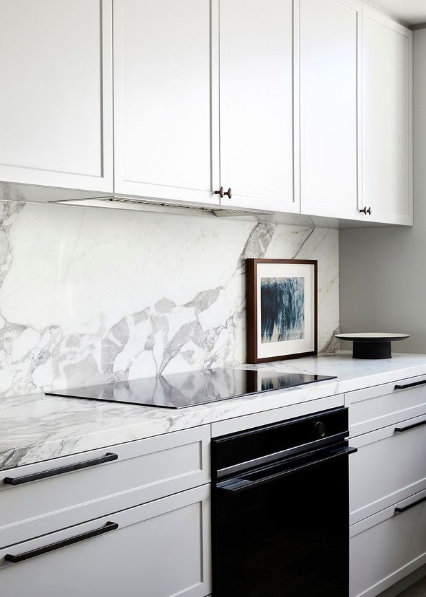 A bright, compact contemporary kitchen featuring white Shaker-style cabinetry with dark linear handles, a dramatic white-and-grey veined stone benchtop and matching full-height splashback, a black glass cooktop and built-in black oven. Minimal decorative styling includes framed artwork and a black bowl on the benchtop.