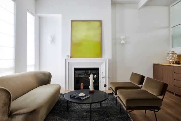 Bright, refined living room with a neutral upholstered sofa, two mid-century style lounge chairs, a round black coffee table, dark textured rug, white fireplace surround, timber flooring, and a low timber sideboard. Large windows and pale walls create an airy, high-contrast interior, with a colourful abstract artwork above the fireplace.