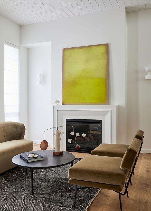Bright, refined living room with a fireplace framed by white decorative moulding, a large lime-yellow artwork above the mantel, white painted plank ceiling, pale walls, timber flooring, and contemporary brown upholstered seating arranged around a round black coffee table. A textured dark rug anchors the seating area.