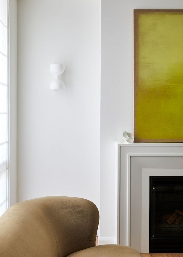 A bright, refined interior detail featuring a white wall with a sculptural wall sconce, a framed lime-green artwork above a white-trimmed fireplace, and a curved tan upholstered armchair in the foreground. Timber flooring and abundant natural light contribute to a warm, contemporary living space.
