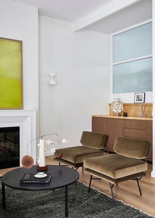 A refined living room with a mid-century-inspired lounge arrangement. Two brown upholstered lounge chairs sit around a round black coffee table over a dark textured rug. The room features a white fireplace surround, pale timber flooring, white painted walls and a white-lined ceiling, with a dark timber sideboard topped by a warm stone or marble surface beneath large textured-glass windows. Decorative lighting, artwork and sculptural objects complete the space.