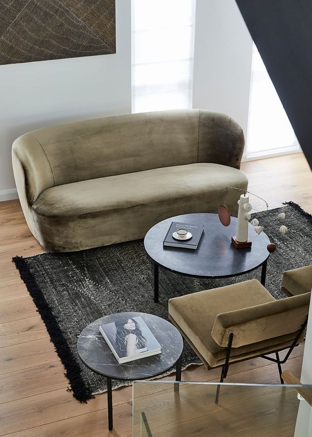 A warmly styled living room featuring a curved taupe velvet sofa, matching upholstered armchair and ottoman, two round black coffee tables, a dark textured area rug, and light timber flooring. Floor-to-ceiling windows with sheer blinds provide abundant natural light, while abstract wall art and understated decorative objects add a refined contemporary feel.