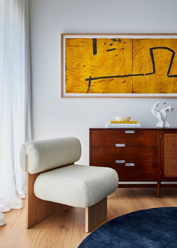 A refined living room corner featuring a sculptural cream boucle lounge chair with a timber frame, a mid-century wooden sideboard with cane-panelled doors, timber flooring, sheer white curtains, and a large abstract ochre artwork in a timber frame. A dark navy round rug and ceramic sculpture add contrast.