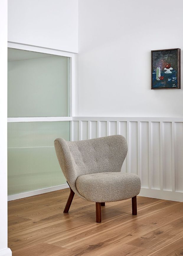 A bright, minimalist sitting area featuring a beige upholstered mid-century-style lounge chair with dark timber legs, positioned on a warm timber floor. White walls with vertical wainscoting and a large frosted-glass window or partition form the backdrop, with a small framed artwork adding a subtle accent.