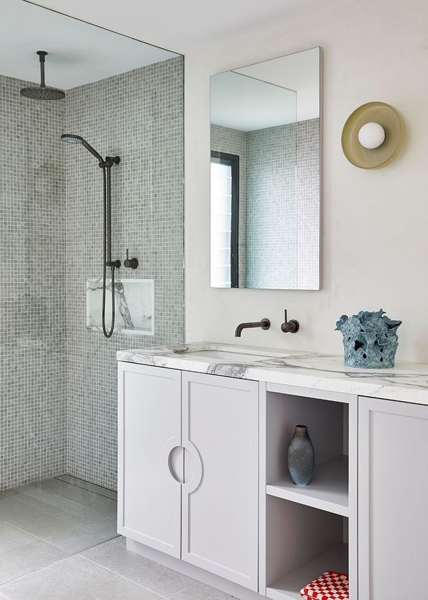 A contemporary bathroom featuring a wall-to-wall glass shower enclosure lined with small pale grey mosaic tiles, dark bronze shower and tapware, and a long white vanity with marble-effect benchtop, paneled cabinetry, open shelving, and round integrated handles. A rectangular mirror and sculptural wall light sit above the vanity. The room has a light, refined palette with subtle decorative accents.