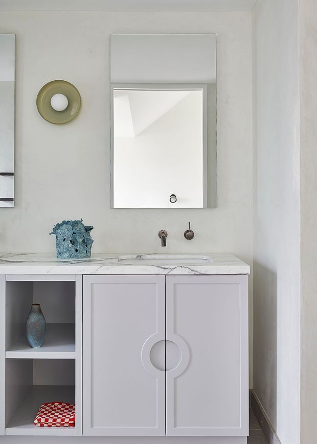 A refined compact bathroom vanity with pale grey built-in cabinetry, a white marble-look benchtop and integrated basin. The cabinetry features decorative geometric circular detailing and open display shelving. Brushed metal wall-mounted tapware sits beneath a simple rectangular mirror, with a sculptural wall sconce and restrained decorative accessories.