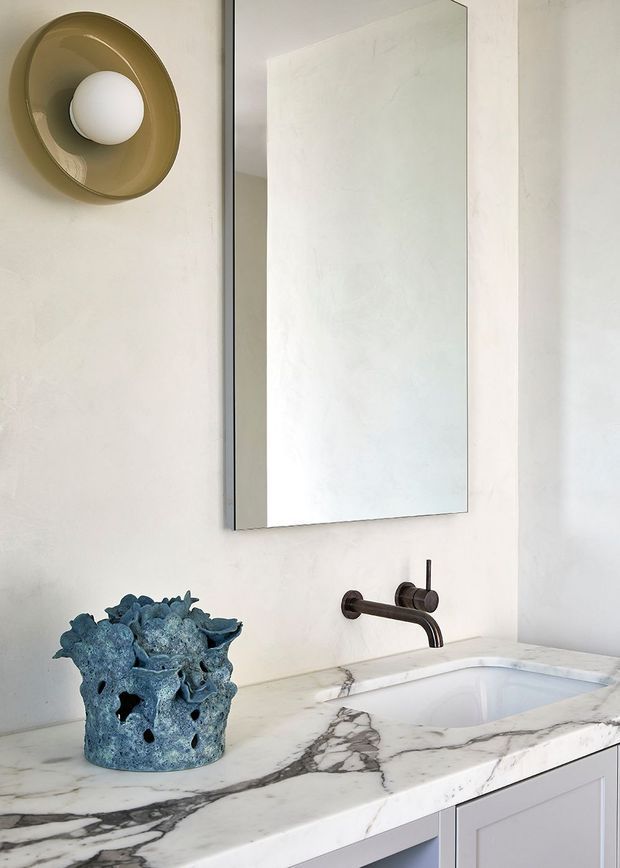 Close-up of a refined bathroom vanity with a white marble countertop featuring bold grey veining, integrated white basin, wall-mounted dark bronze faucet, large frameless rectangular mirror, and sculptural bronze wall light. A blue textured decorative vessel adds contrast against the pale plaster walls and light cabinetry.