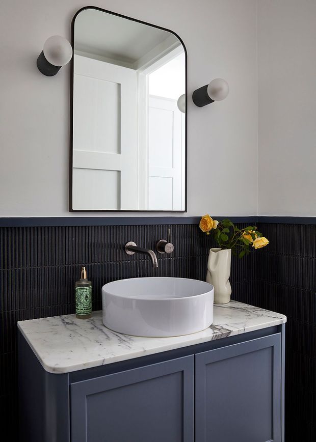 A compact, refined bathroom vanity area featuring a dark navy-blue fluted wall finish and matching painted vanity cabinetry, topped with a white veined marble benchtop and a white round vessel basin. A black-framed arched rectangular mirror is flanked by globe wall sconces, with brushed metal tapware and a small floral arrangement adding detail.