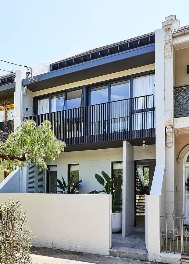 Street-facing view of a contemporary multi-storey townhouse or terrace extension, with a dark metal balcony railing, large glazed doors, pale rendered and painted masonry walls, and a flat roof with a raised parapet detail. A landscaped entry passage and courtyard planting sit beside an adjoining ornate heritage facade.