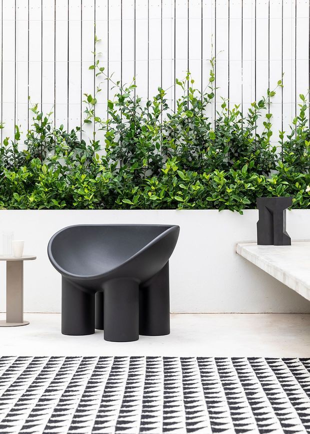 A refined contemporary interior vignette featuring a sculptural matte-black lounge chair with rounded bowl-like form and cylindrical legs, positioned against a white vertical-panel wall. A dense green planting bed runs along the wall, complemented by a pale stone or plaster bench and a small black sculptural object. A black-and-white geometric rug spans the foreground, with a minimalist side table at left.
